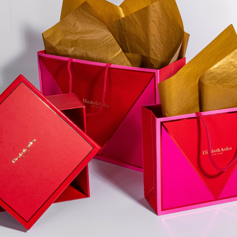 Red and pink Elizabeth Arden gift bags and boxes from their exquisite collections are displayed with gold tissue paper peeking out. The bags, adorned with rope handles, proudly feature the brand's name in gold on each item, set against a plain white surface.