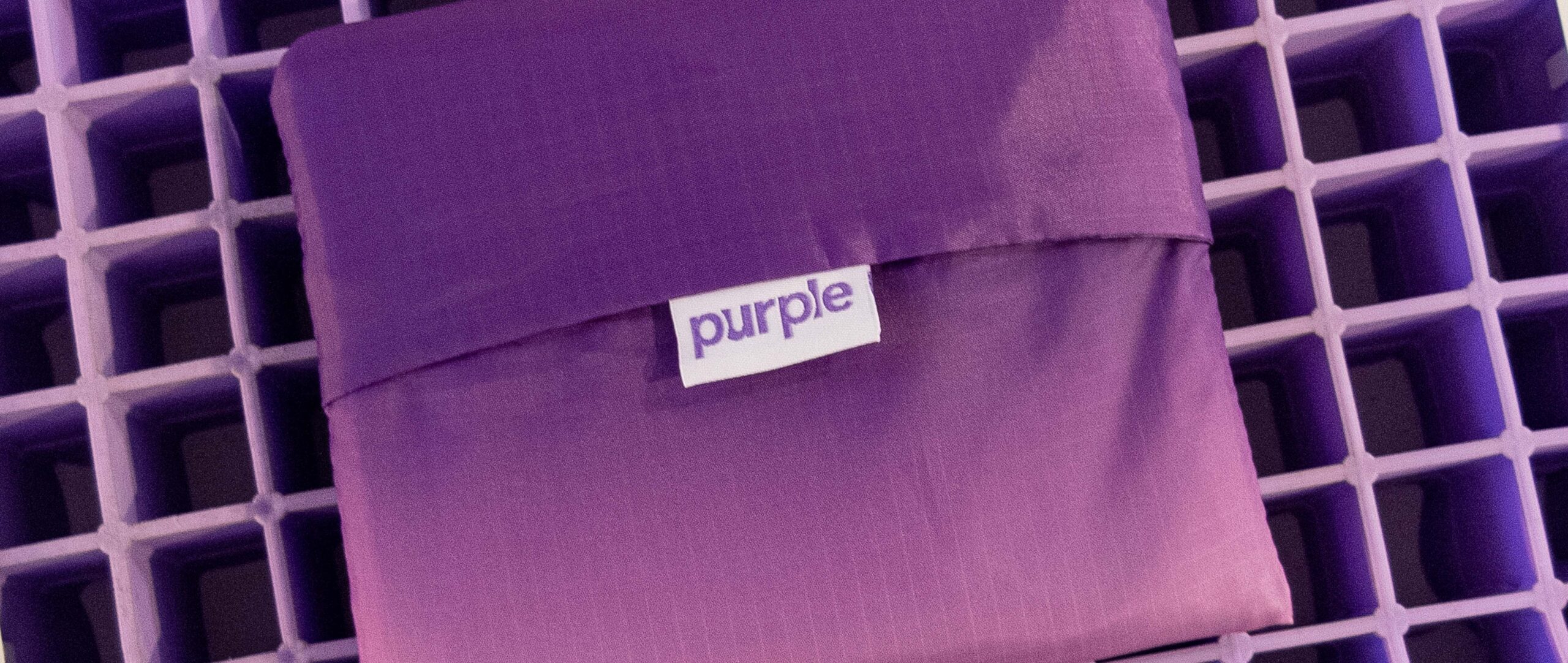 A purple fabric pouch sits on a waffle-like purple surface, showcasing a small white label with the word purple. This pouch is perfect for storing your items inside a matching XL tote, combining style and functionality seamlessly.