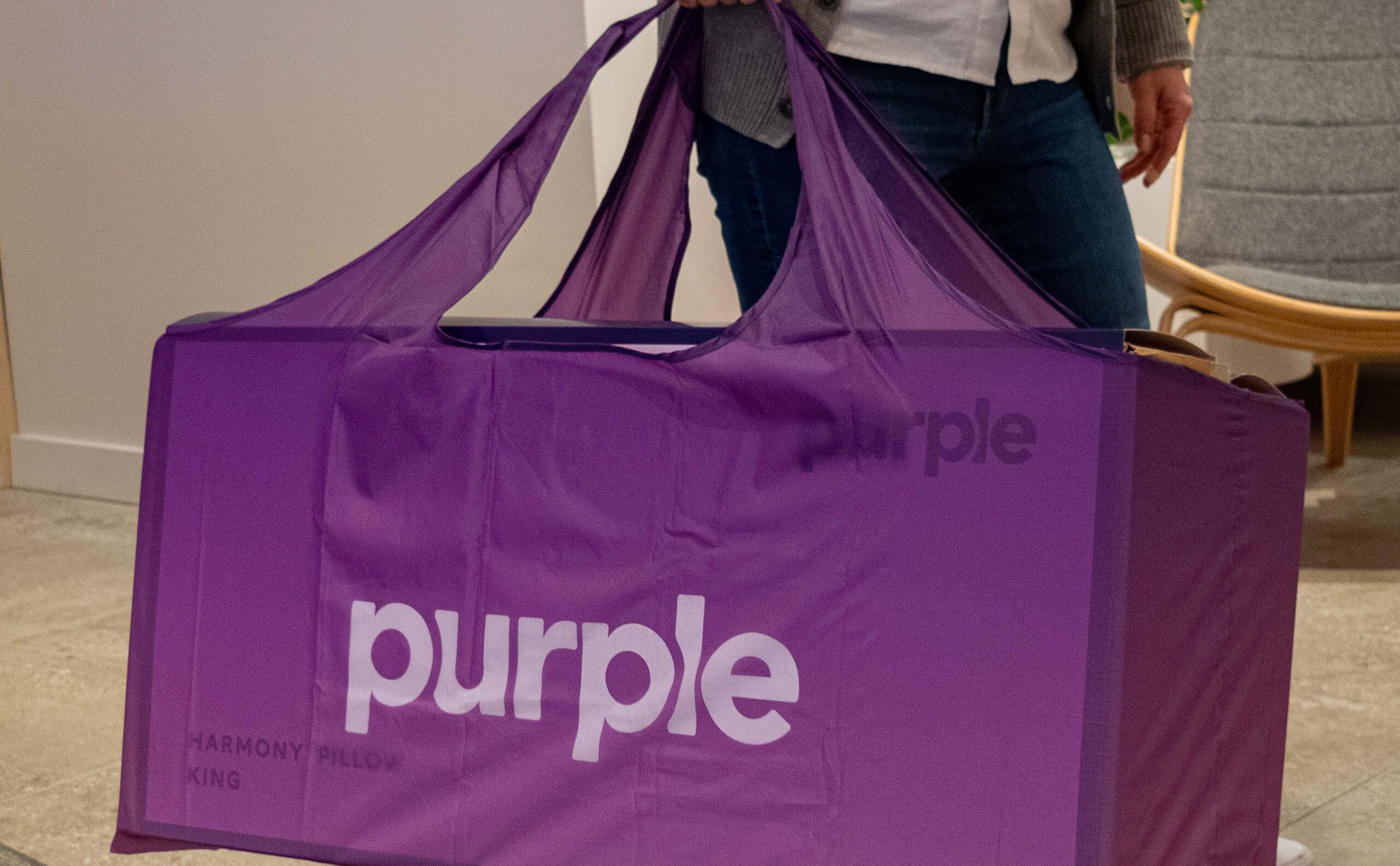 A person carrying a large, rectangular purple reusable tote with the brand name Purple printed on it. The tote appears to contain a Harmony Pillow, King size. The person is wearing a gray cardigan and jeans, standing indoors.