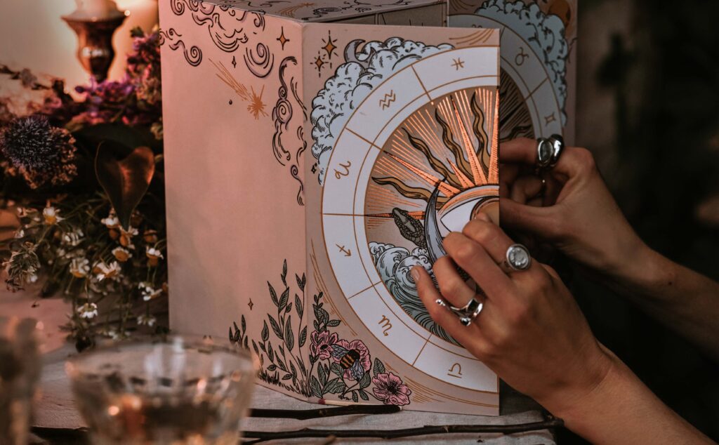 A person with rings is opening a book featuring a zodiac wheel illustration, nestled within an Advent Box surrounded by flowers and candles. The scene exudes a mystical and vintage ambiance, hinting at the magic to come in AWE 2024.