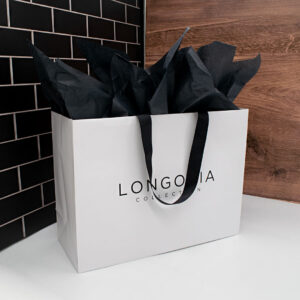 A white shopping bag with black handles, labeled Longoria Collection, brimming with black tissue paper, exudes a high-end shopping vibe. It rests elegantly against a backdrop of black tiles and wooden paneling.